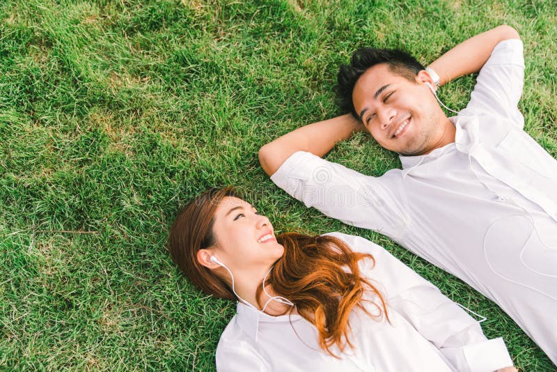Giovani coppie o studenti di college adorabili asiatici che si riposano insieme sull'erba, ascoltando la musica, vista superiore immagine stock Giovani coppie o studenti di college adorabili asiatici che si riposano insieme sull'erba, ascoltando la musica, vista superiore immagine stock