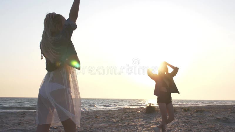 Giovani donne del movimento lento vestite in rivestimenti del denim divertendosi sulla spiaggia nella sera nei raggi del tramonto