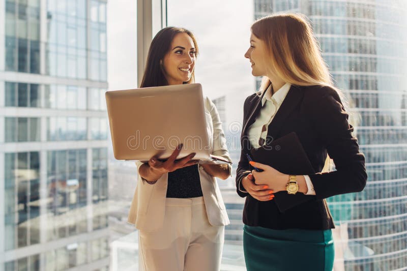 Giovani donne sorridenti con il computer portatile che sta e che parla nell'ufficio fotografia stock Giovani donne sorridenti con il computer portatile che sta e che parla nell'ufficio fotografia stock