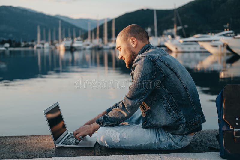 Giovani free lance dell'uomo dei pantaloni a vita bassa che lavorano con il computer portatile che si siede vicino al Se immagini stock Giovani free lance dell'uomo dei pantaloni a vita bassa che lavorano con il computer portatile che si siede vicino al Se immagini stock