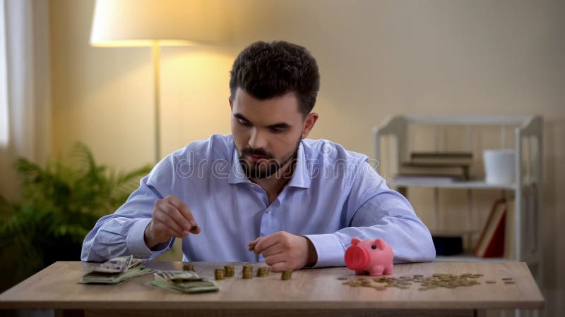 Giovani monete di conteggio maschii, porcellino salvadanaio che fa una pausa, alfabetizzazione finanziaria, bilancio immagine stock libera da diritti Giovani monete di conteggio maschii, porcellino salvadanaio che fa una pausa, alfabetizzazione finanziaria, bilancio immagine stock libera da diritti
