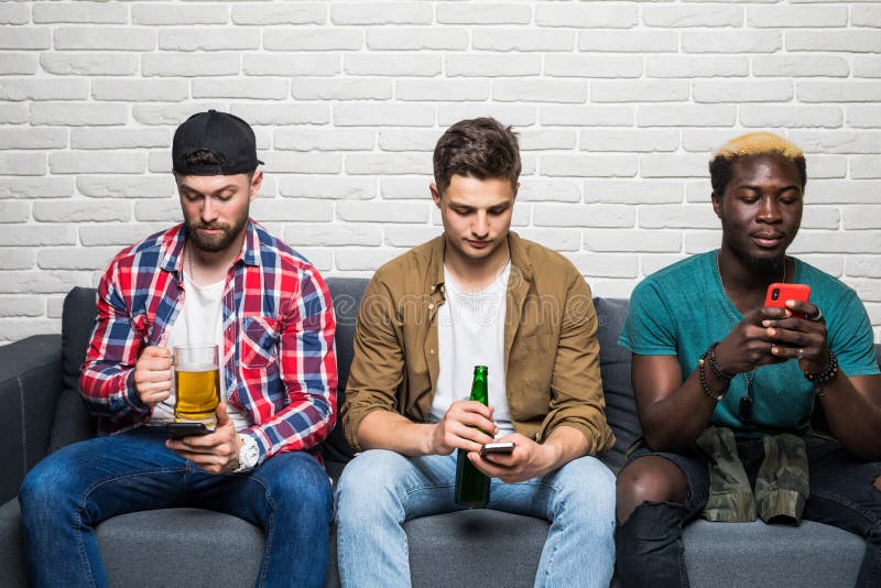 Giovani uomini attraenti della corsa mista che vanno in giro a casa, birra bevente e mangianti pizza mentre per mezzo dei loro sm immagine stock Giovani uomini attraenti della corsa mista che vanno in giro a casa, birra bevente e mangianti pizza mentre per mezzo dei loro sm immagine stock