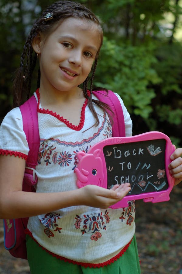 Cute young girl with rucksack holding black or chalkboard with back to school text; trees in background. Cute young girl with rucksack holding black or chalkboard with back to school text; trees in background.