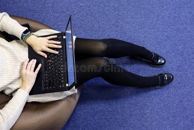 Girl in black pantyhose and knitted sweater sitting on padded stool with laptop on her knees, top view