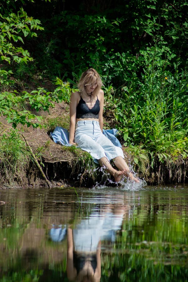 A Girl with Blond Hair on the Lake Shore Has Her Feet in the Water and ...