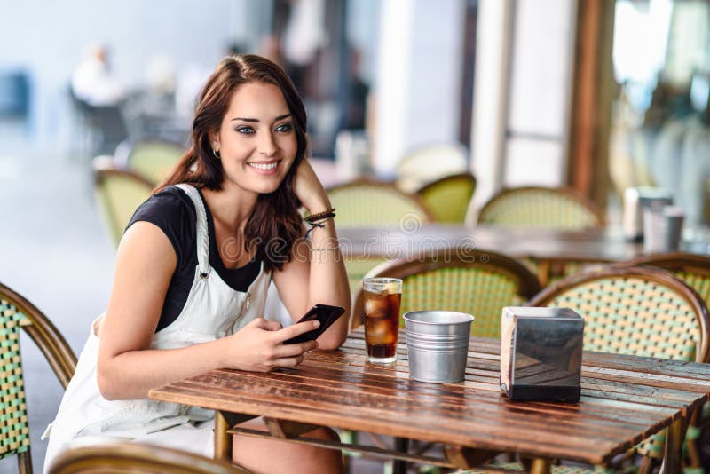 Girl with blue eyes sitting on urban cafe using smart phone smiling