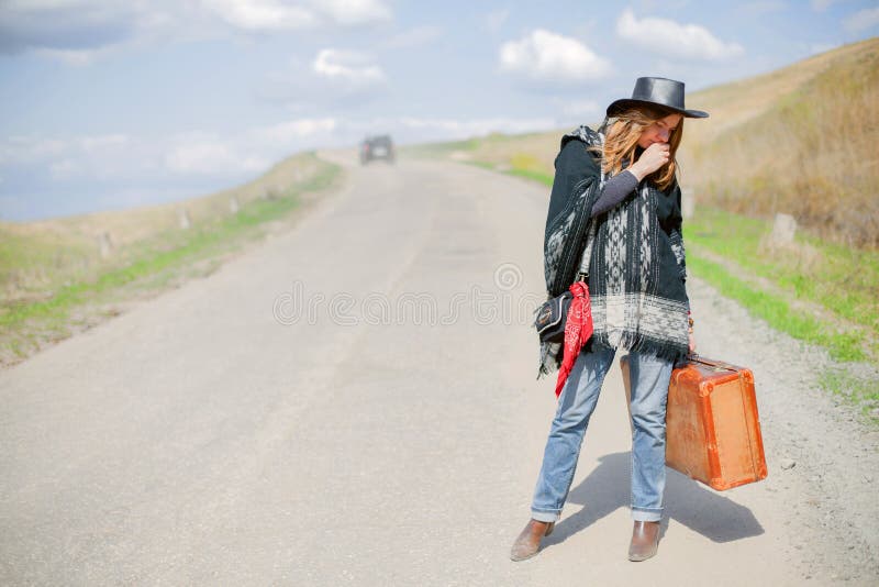 A girl in blue jeans, a poncho, a black leather hat with an old brown suitcase in her hands is on the road. stock images A girl in blue jeans, a poncho, a black leather hat with an old brown suitcase in her hands is on the road. stock images
