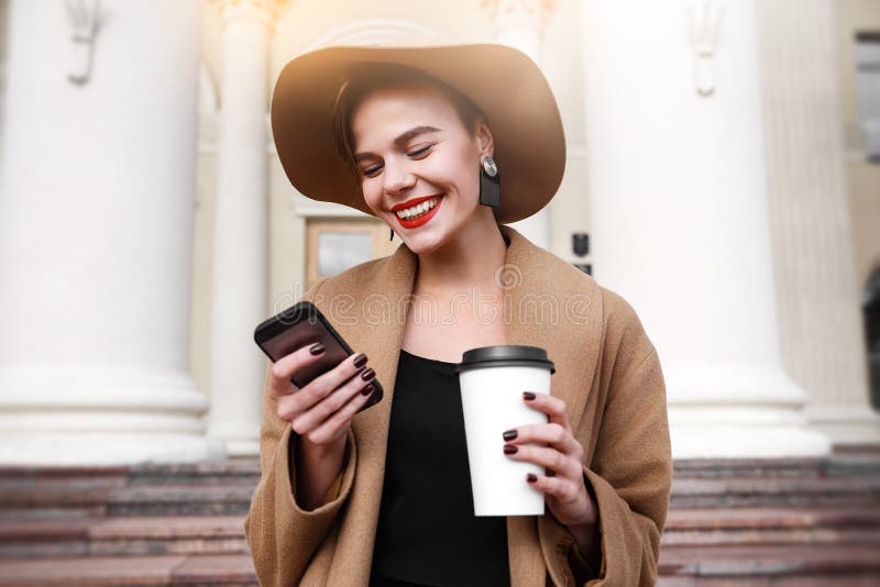 Girl in a brown coat a brown hat is walking and posing in the city interiors. The girl is smiling, checking her