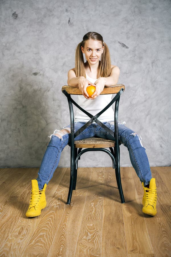Pretty girl with ponytails holding orange and sitting on a chair backwards.