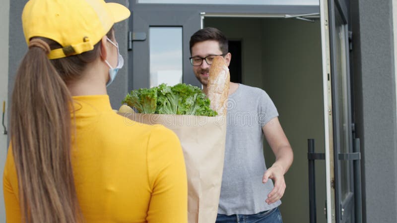 Girl Delivery Service Worker Brings Groceries. She Is Met By Young Man And Takes Package. In Package Are Bread And Salad. Girl Wears Mask And Gloves To Protect