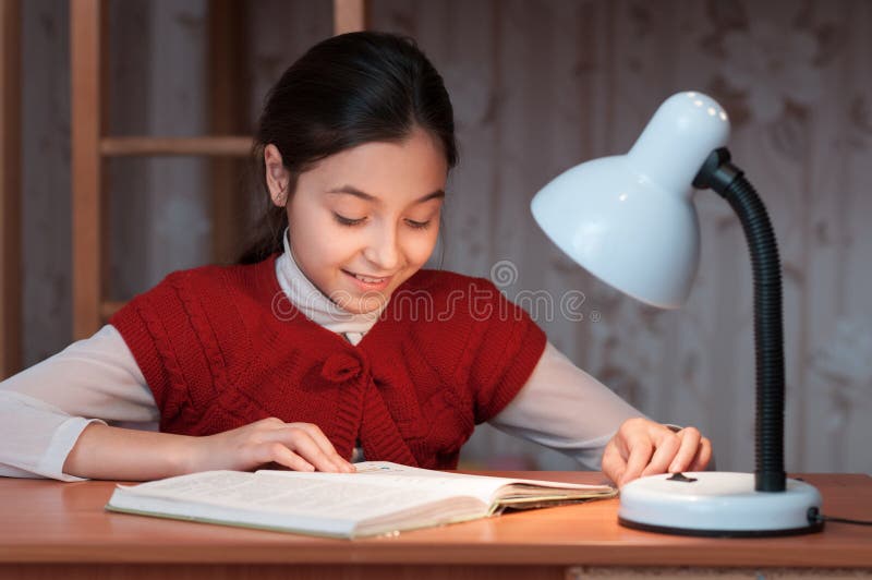 Girl at desk reading a book by light of lamp