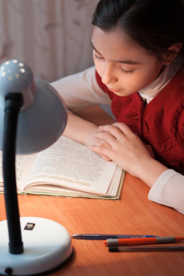 Girl at desk reading a book by light of the lamp