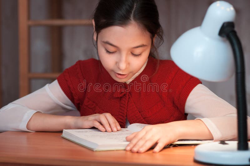 Girl at desk reading a book by light of the lamp