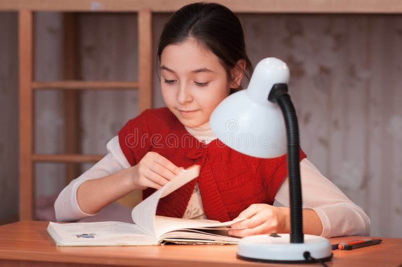 Girl at desk reading a book by light of the lamp