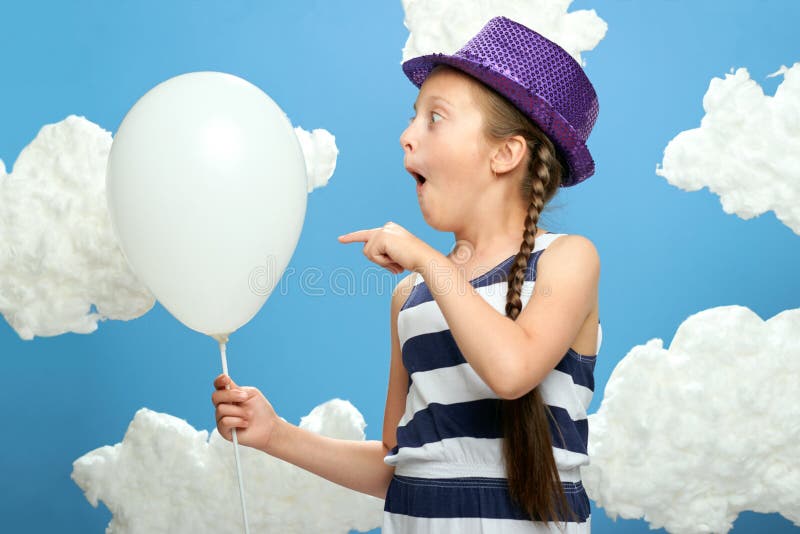 Girl dressed in striped dress and color hat posing on a blue background with cotton clouds, white air balloon, the concept of summ
