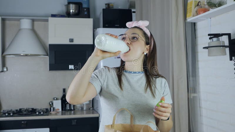 The girl drinks milk and unpacks a paper bag with fruit in the kitchen after shopping