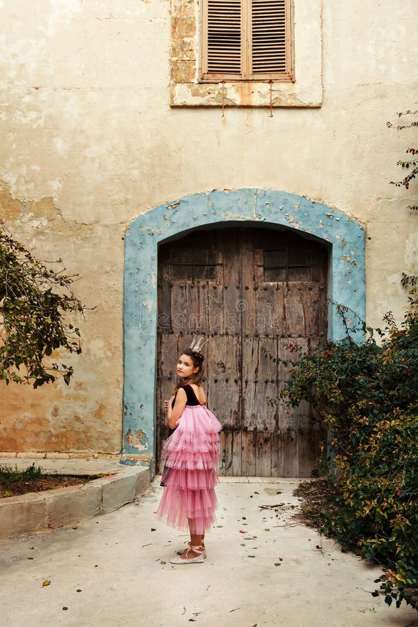 A girl in a fairy dress of a princess with a crown turns around walkng to the door of an ancient castle house