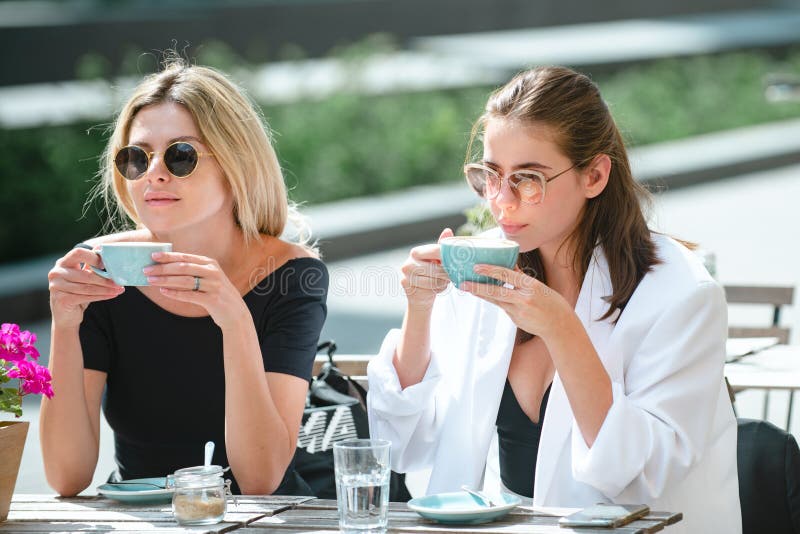 Girl friends in cafe outdoor. Two beautiful girls with cups coffee in summer cafe. Outdoors portrait of two young