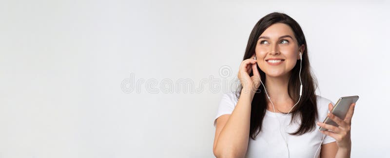 Smiling Girl Enjoying Music In Earphones On Phone Listening To Favorite Song Standing On White Background. Panorama, Free Space. Smiling Girl Enjoying Music In Earphones On Phone Listening To Favorite Song Standing On White Background. Panorama, Free Space
