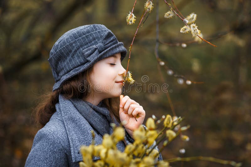 A girl in grey coat and a cute gray hat in the forest in early spring with a willow branch of twigs smelling twigs.