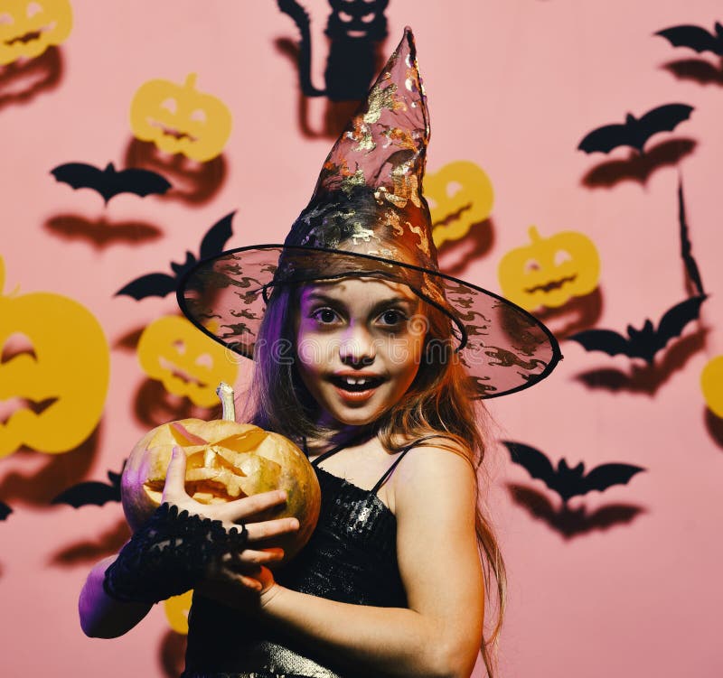 Girl with happy face on pink background with bats, pumpkins. Girl with happy face on pink background with bats and pumpkins decor. Little witch wearing black hat stock photo Girl with happy face on pink background with bats, pumpkins. Girl with happy face on pink background with bats and pumpkins decor. Little witch wearing black hat stock photo