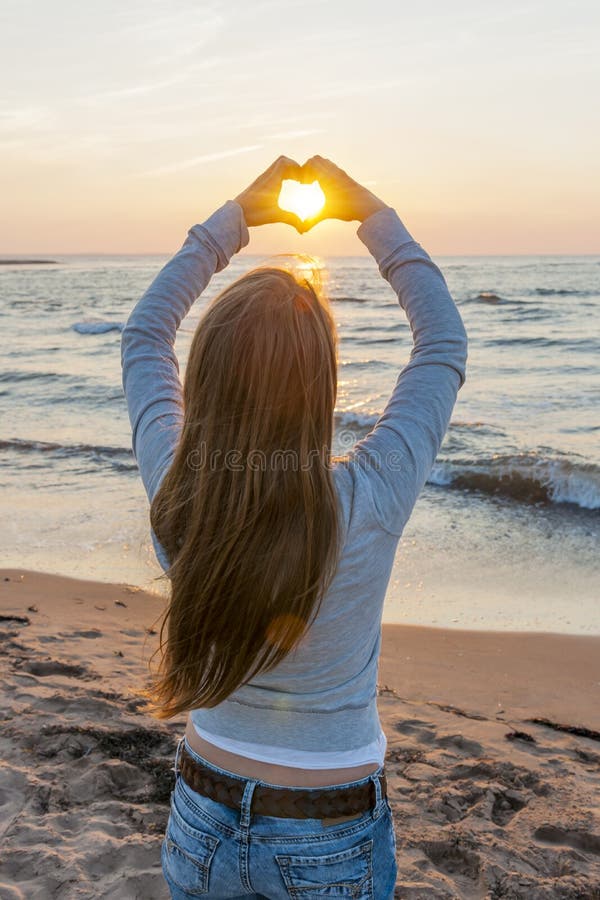 Blonde young girl holding hands in heart shape framing setting sun at sunset on ocean beach. Blonde young girl holding hands in heart shape framing setting sun at sunset on ocean beach