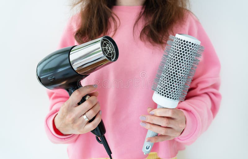 The girl holds a hair dryer and a brush in her hands. Drying long brown hair with a round brush for styling unruly hair. Beauty