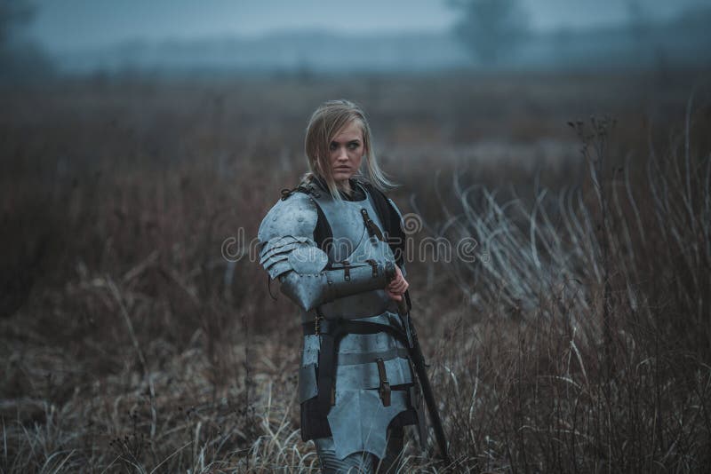 Girl in image of Jeanne d`Arc in armor and with sword in her hands stands on meadow. Back view. royalty free stock image Girl in image of Jeanne d`Arc in armor and with sword in her hands stands on meadow. Back view. royalty free stock image