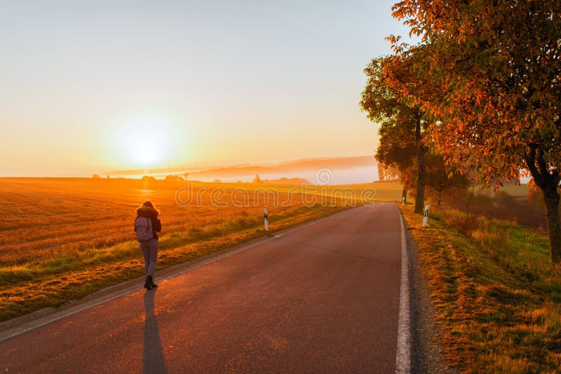 A girl in a jacket with a backpack and suitcase on the road against the background of the autumn field and sunset