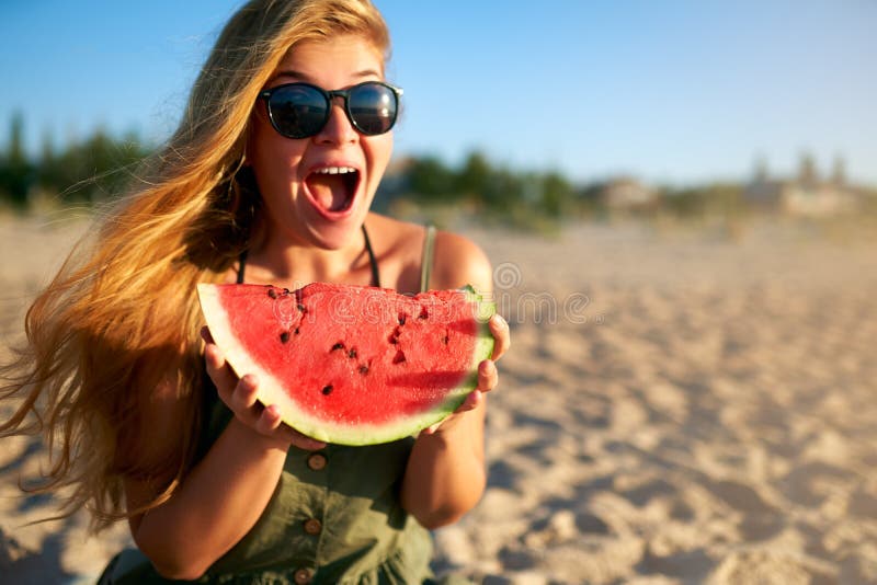 Happy young woman in glasses eating watermelon on the sandy beach on vacation. Girl joyfully holding fresh watermelon