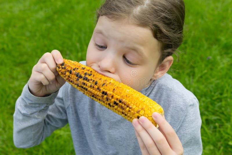 The girl keeps hands an ear of corn and bites with appetite, against the green lawn stock photography The girl keeps hands an ear of corn and bites with appetite, against the green lawn stock photography