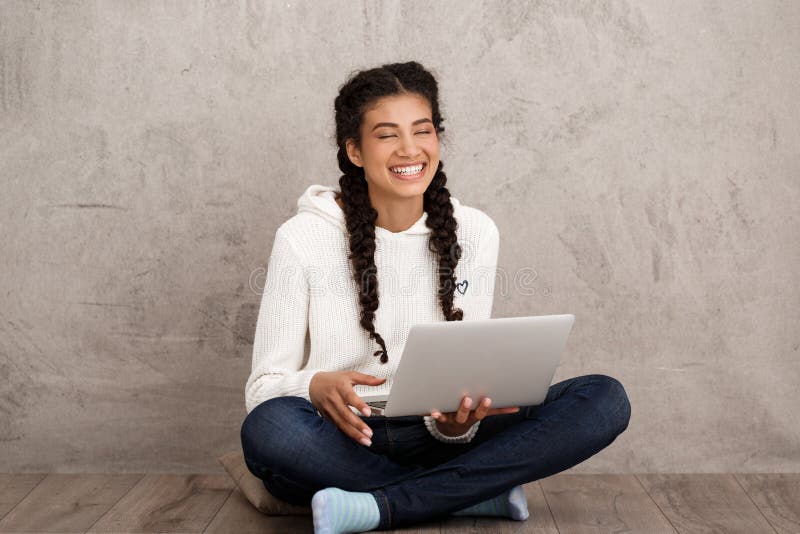 Girl laughing, holding laptop, sitting on floor over beige background.