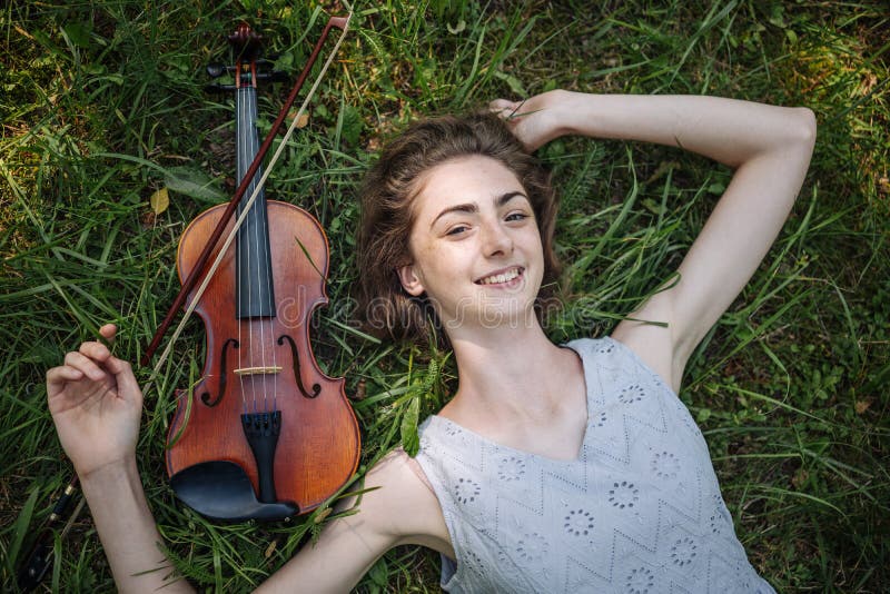 The girl lies with a violin on the grass in a city park.