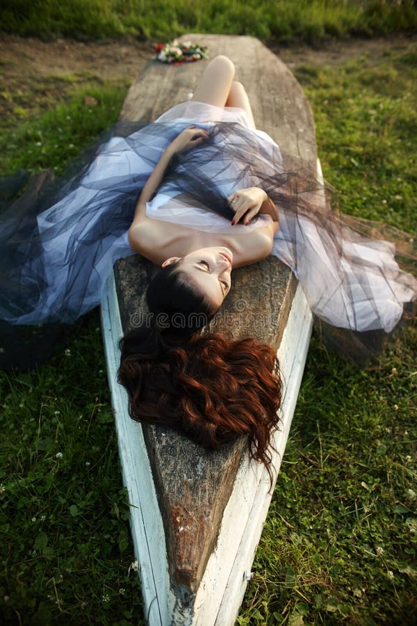 Girl in a long dress near the lake sits on an old boat, a beautiful woman in the rays of the evening sun in the autumn in the