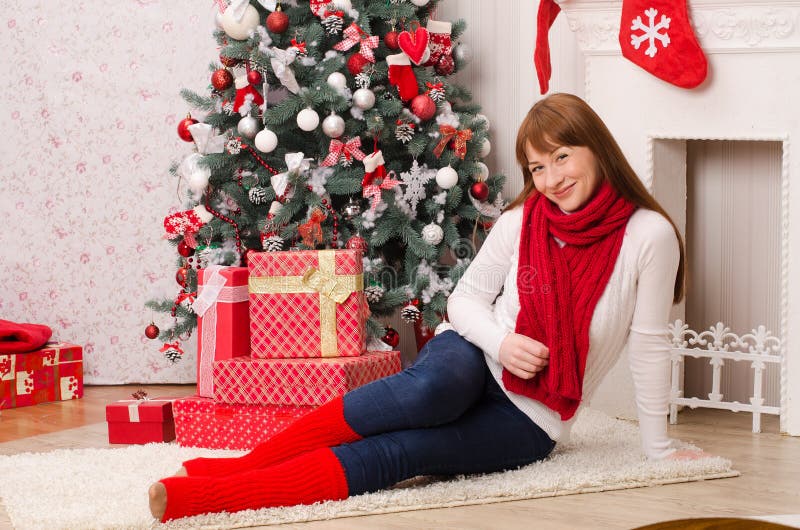 Girl with long hair in a white sweater, and a red scarf and golfs, sits near a Christmas tree and boxes with gifts