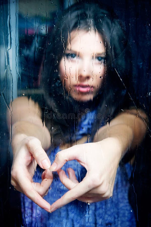 Girl making heart from fingers behind wet window