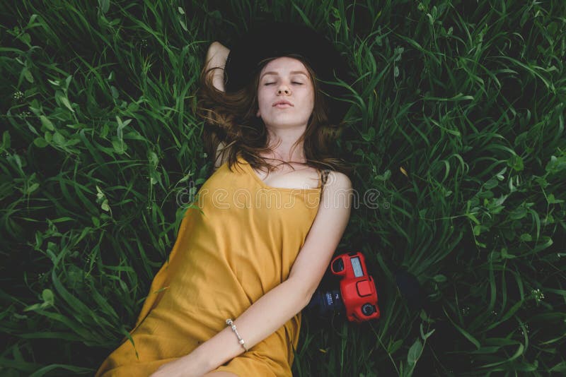 A girl in a mustard-colored dress and a hat lies on a green field next to a red camera with her eyes closed