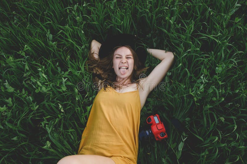 A girl in a mustard-colored dress and a hat lies on a green field next to a red camera and shows her tongue