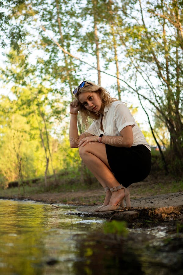 A Blonde Woman in a White Shirt Poses on the Bank of a River or Lake ...