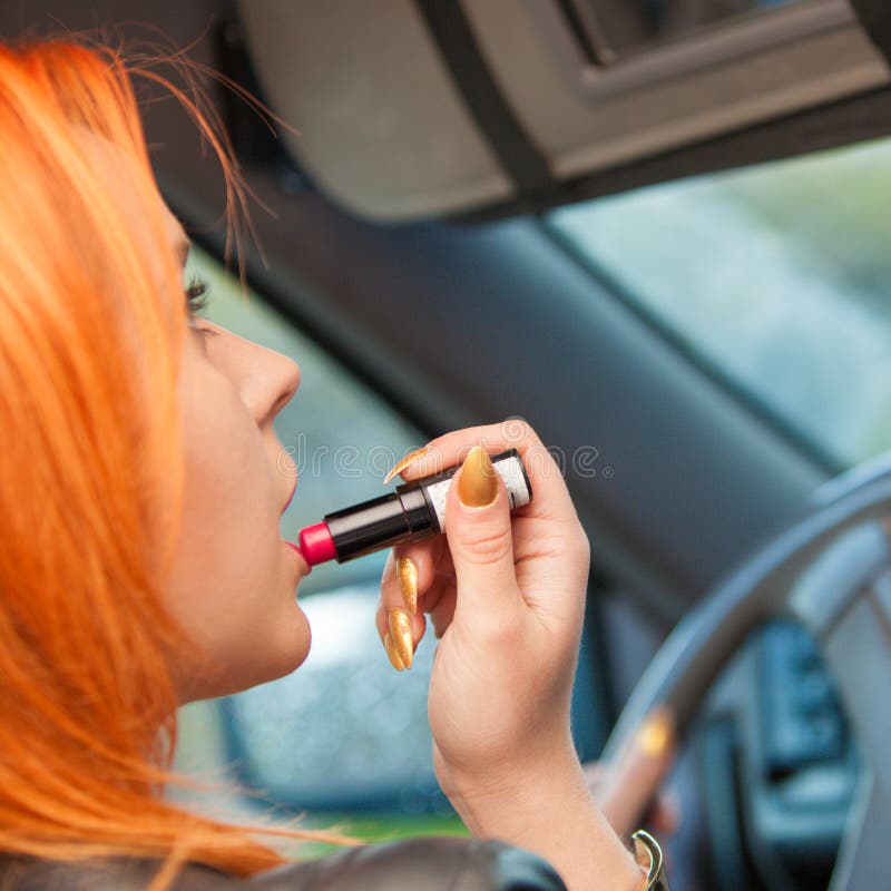 Concept of danger driving. Young woman driver red haired teenage girl painting her lips doing applying make up while driving the car. Concept of danger driving. Young woman driver red haired teenage girl painting her lips doing applying make up while driving the car.