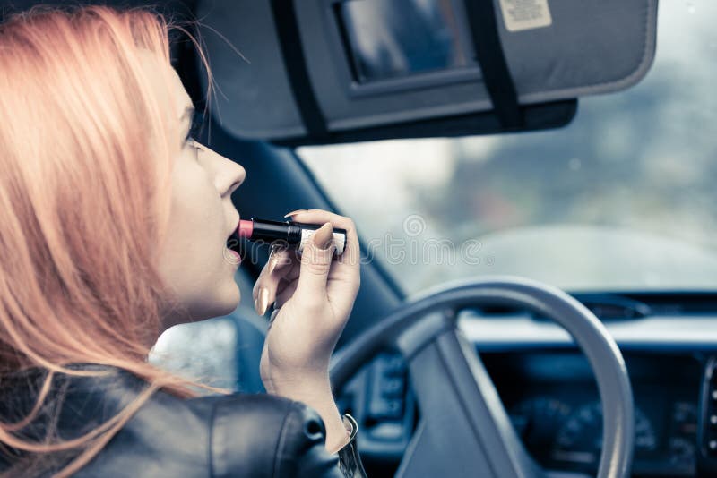 Concept of danger driving. Young woman driver red haired teenage girl painting her lips doing applying make up while driving the car. Concept of danger driving. Young woman driver red haired teenage girl painting her lips doing applying make up while driving the car.