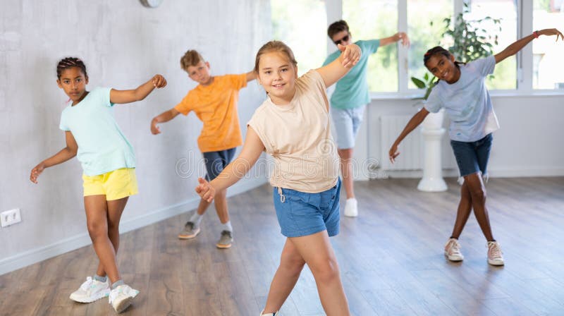 Female child performs choreographic exercises and teaches energetic mobile social dance waacking together with friends. Young girls and guys repeat movements, train in spacious studio unfocused. Female child performs choreographic exercises and teaches energetic mobile social dance waacking together with friends. Young girls and guys repeat movements, train in spacious studio unfocused