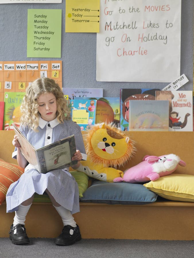 Elementary schoolgirl reading book on couch in classroom