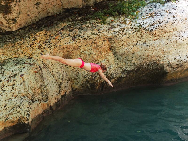 A girl in a red swimsuit dives from a low cliff into the river on a summer day