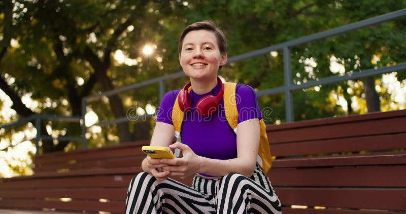 A girl with a short haircut in a purple top, striped pants and a yellow backpack sits on brown stands and chats on a