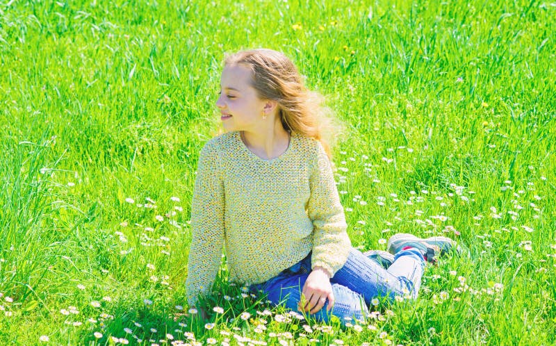 Girl sits on grass at grassplot, green background. Child enjoy spring sunny weather while sitting at meadow. Springtime