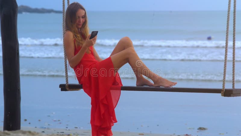 Girl Sits on Swing Chats on Phone against Wavesurf