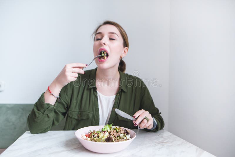 The girl sits at the table and eats a salad with a fork. The girl bites green salad and gets plucked. Eating a healthy meal