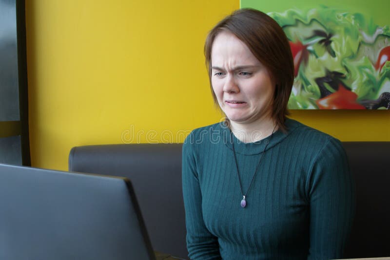 A girl sits, works at a laptop at a table in a cafe on a brown sofa against a yellow wall. On the face is an expression