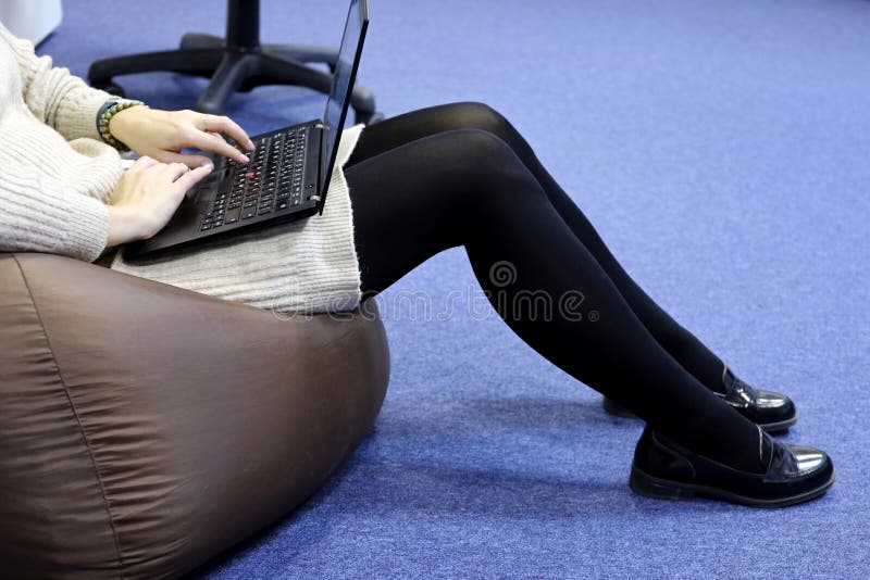 Girl in black pantyhose and knitted sweater sitting on padded stool with laptop on her knees, side view